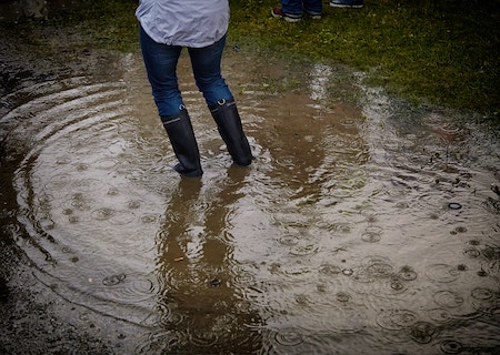 legs of person wearing blue jeans and black rain boots standing in a brown puddle